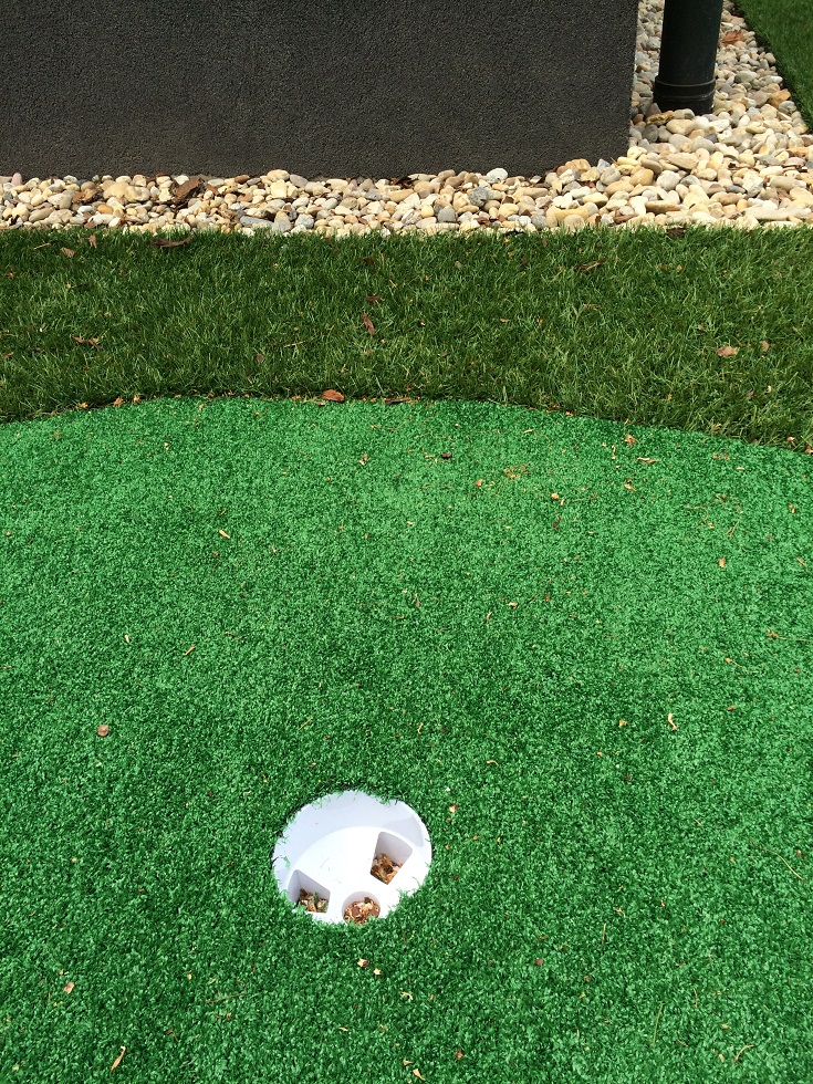 Műfüves golfpálya golflukkal és kaviccsal szegélyezett fekete fallal a háttérben.